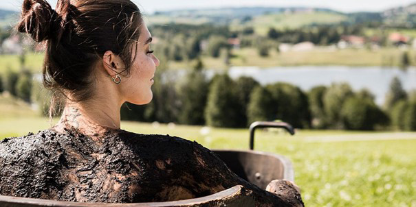 Ein Moorbad einer jungen Frau in einer Holztonnen im Freien mit der Sicht an den Soier See der im Hintergrund zu sehen ist