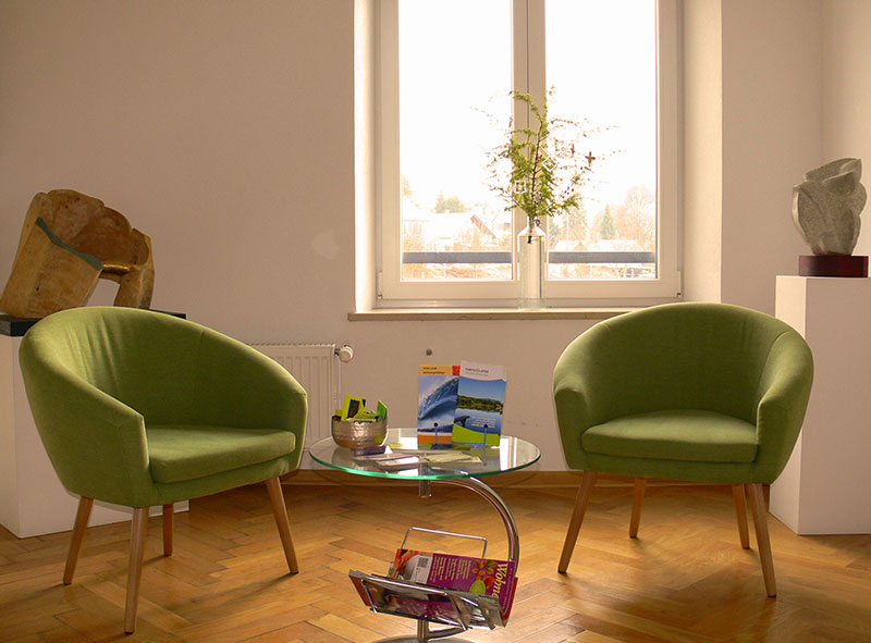 Vita Dr. med. Reingard Herbst - Wartebereich der Arztpraxis ist hell mit 2 grünen Stoffsesseln, ein Holzboden in Fischgrätmuster und ein Fenster